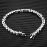 White gold plated cubic zirconia tennis chain bracelet laid open on black ribbed surface, box clasp and tongue detail visible, iced out tennis bracelet product detail